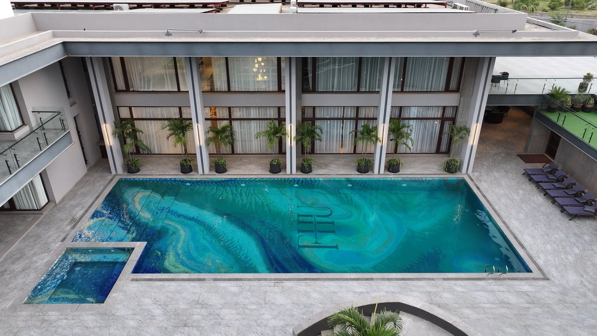 Aerial view of the main swimming pool with the FHU Resorts monogram set into its agate-swirl mosaic floor, framed by the glass villa facade and lined palms at FHU Resorts Karachi